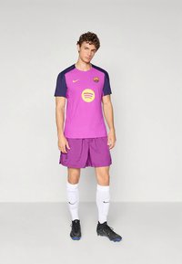 Vīrietis tērpies violeta un tumši zilā futbola formā ar dzelteniem logotipiem, baltās zeķēs un melniem futbola apaviem, stāvot pret vienkāršu fonu.