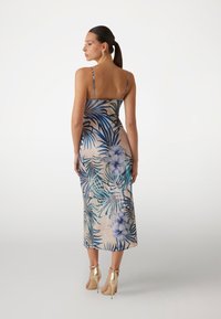 Abito maxi floreale con spalline sottili, caratterizzato da uno sfondo beige, foglie tropicali blu e fiori blu chiaro. Tessuto liscio, design aderente.