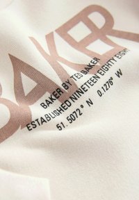 Weißer Stoff mit einer weicheren Textur, der einen großen braunen "BAKER"-Druck und kleinere schwarze Schriftzüge mit Koordinaten und Markeninformationen zeigt.