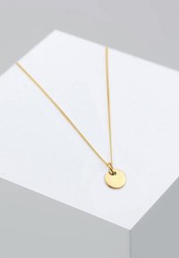 Collana con ciondolo in oro, con un disco rotondo spazzolato appeso a una catena sottile e delicata; esposto su uno sfondo geometrico grigio chiaro.