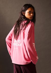 Femeie cu păr lung și închis la culoare, îmbrăcată cu o bluză roz cu mâneci lungi și lejere, având pe spate un text alb "dare to standout", întoarsă lateral pe un fundal întunecat.