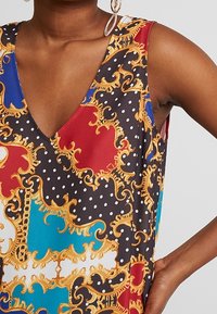 Färgglad ärmlös topp med en djup V-ringning, prydd med ett djärvt mönster av röd, blå och svart med guldaccenter och prickar på ett silkesliknande tyg.