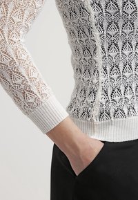 Bras portant un pull en maille blanche à dentelle avec poignet, main partiellement glissée dans la poche d'un pantalon noir sur un fond uni.