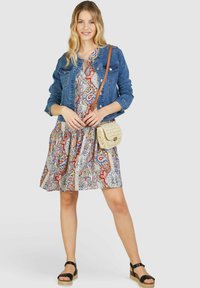 Džínová bunda přes barevné vzorované šaty s červenými, modrými a bílými odstíny. Modelka drží tkanou crossbody tašku; černé sandály dokončují outfit.