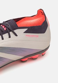 Botte de football grise avec des accents noirs et des détails roses. Comprend un matériau texturé, un design à coupe basse et une semelle extérieure à crampons pour l'adhérence.