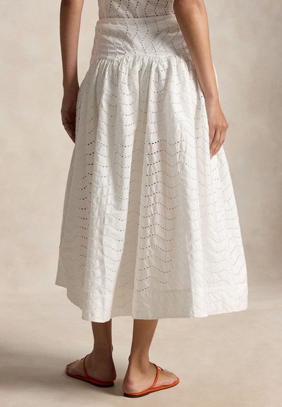 Γυναίκα με λευκή μίντι φούστα με κυματιστά μοτίβα με οπές και πορτοκαλί επίπεδα σανδάλια, στέκεται μπροστά σε ουδέτερο φόντο.