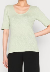 Maglietta a maniche corte verde chiaro con scollatura a barca, realizzata in tessuto morbido e testurizzato. Indossata con pantaloni neri, che mettono in risalto una forma aderente.