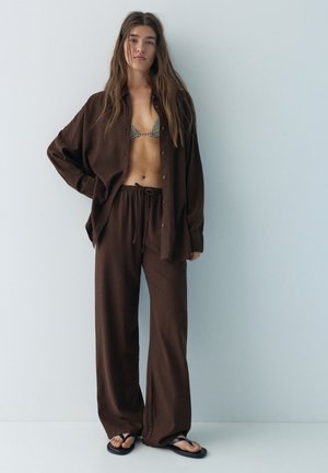 Kvinde iført løstsiddende brune bukser, matchende oversized skjorte, ternet bikini top og sorte flip-flops, stående mod en ensfarvet lys væg.