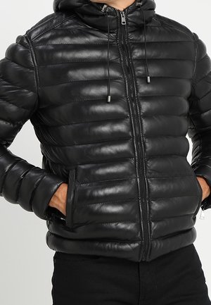 Doudoune matelassée noire avec capuche, fermeture éclair à l'avant, poches latérales et cordons, portée par une personne avec les mains dans les poches.