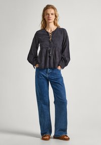 Blusa grigio scuro con scollatura allacciata, orlo arricciato e maniche lunghe a palloncino, abbinata a jeans a gamba larga blu e sandali marroni.