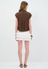 Camicia corta a maniche corte marrone a costine con colletto, abbinata a una mini gonna bianca a strati con volant. Modella di spalle rispetto alla telecamera.