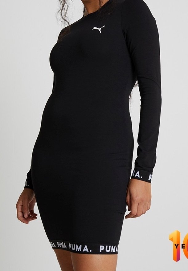 Femme portant une robe noire ajustée à manches longues de Puma avec des logos Puma blancs sur la poitrine, les poignets et l'ourlet, sur un fond uni.