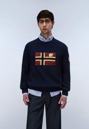 Ung mand iført en marineblå striktrøje med et rød-hvidt flagmærke og tallet 87, over en lyseblå skjorte og mørke jeans, står stille.