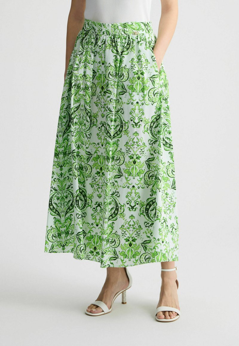 Falda maxi hecha de una tela ligera, con un estampado floral y de paisley en verde, con una cinturilla elástica y bolsillos laterales. Combinada con tacones blancos.