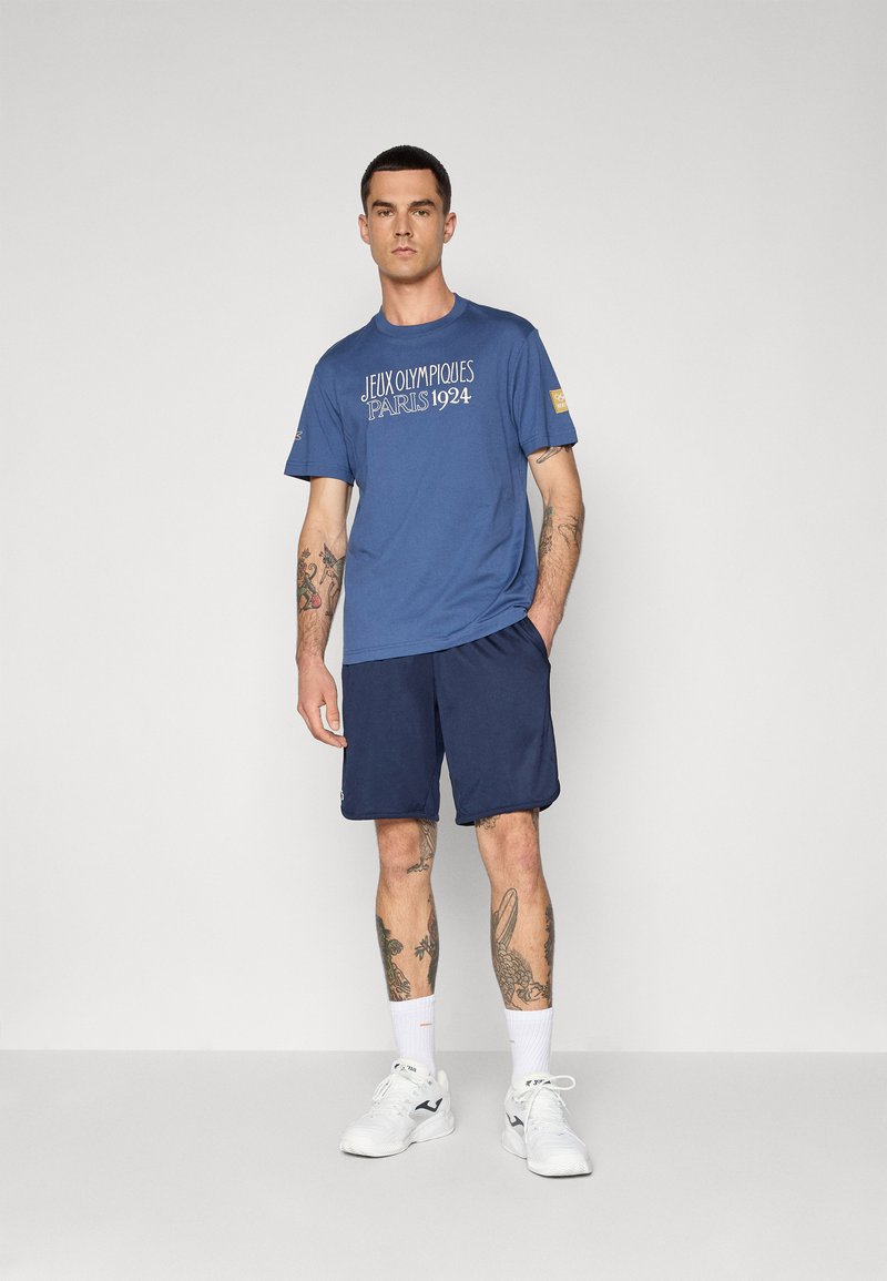 T-shirt en coton bleu avec le texte blanc "JEUX OLYMPIQUES PARIS 1924," assorti à un short marine et des chaussures de sport blanches.
