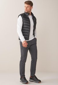 Sort puffer vest, hvid langærmet skjorte, grå tapered bukser med lynlåse og sorte sneakers med orange detaljer, stående mod en neutral baggrund.