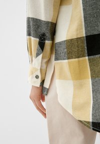 Camicia flanella a quadri con quadrati neri, gialli e bianchi, dotata di polsini con bottone e un tessuto testurizzato. L'orlo è curvo.