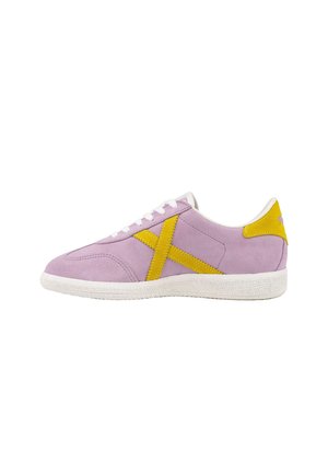 Zapatilla deportiva de ante morada con una franja amarilla en forma de X y lengüeta en el talón, cordones blancos y suela blanca texturizada, mostrada de lado sobre un fondo blanco.