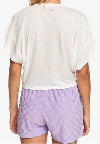 Weißes Kurzarmoberteil mit abgerundetem Saum und kleinem Logo auf dem Rücken, kombiniert mit lavendelnen, strukturierten Karoshorts.