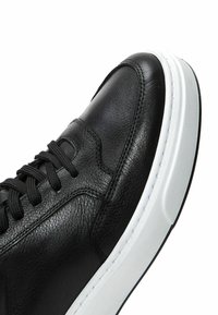 Zapatilla de cuero negro con textura lisa, que presenta una entresuela blanca, cordones negros y detalles de costura en contraste.
