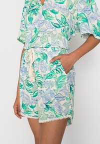 Macacão com padrão floral em tecido leve, apresentando folhas verdes e azuis, bolsos laterais, cintura com cordão de ajuste e acabamento em renda decorativa.