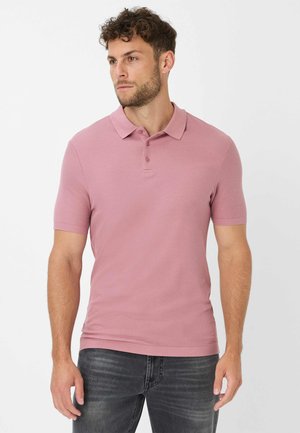 Short-sleeve polo shirt in light pink, featuring a collar, three-button placket, and textured fabric. Paired with dark denim jeans.