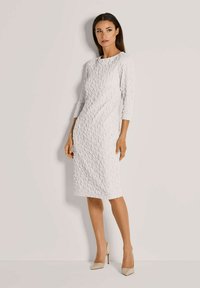 Weißes Kleid mit texturiertem, blattähnlichem Muster, dreiviertel Ärmel und knielanger Schnitt. Getragen mit hautfarbenen High Heels.
