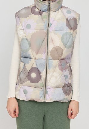 Veste matelassée beige sans manches avec motifs floraux pastel et fermeture éclair à l’avant, portée sur un haut blanc à manches longues et un pantalon vert.