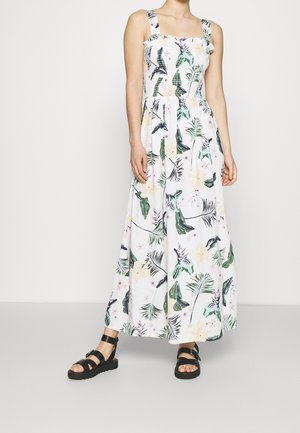 Femme portant une robe blanche sans manches, longue jusqu'aux chevilles, avec des imprimés de feuilles et de fleurs tropicales vertes et jaunes, et des sandales noires.