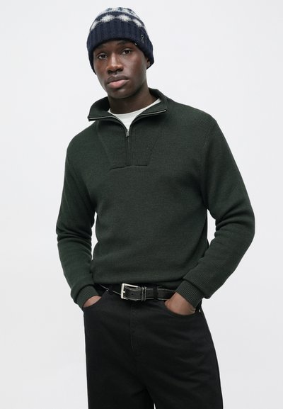 Σκούρο πράσινο πλεκτό πουλόβερ με γιακά που κλείνει με φερμουάρ, συνδυασμένο με μαύρο παντελόνι και σκουρόχρωμο καπέλο με άσπρα σχέδια. Απλή ζώνη ως διακοσμητικό στοιχείο.