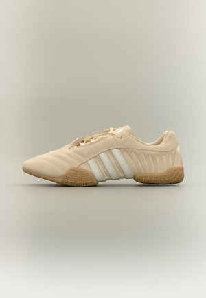 Zapatilla beige de ante con franjas blancas y suela de goma texturizada, vista lateral sobre fondo neutro.