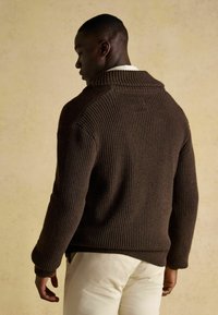 Barna kötött pulóver bordázott textúrával, széles gallérral és leejtett vállakkal, világos színű nadrággal párosítva. Diszkrét kézműves részletek láthatók.