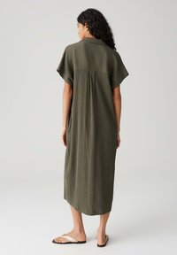 Olive green midi dress with short sleeves, textured fabric, and back pleat detail, paired with flat beige sandals.