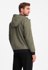 Giacca con cappuccio verde oliva realizzata in tessuto liscio, con polsini ed elastico in vita; presenta una vestibilità rilassata. Vista da dietro.