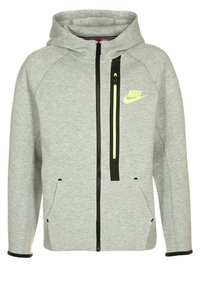 Grijs gewatteerd jack van zachte stof. Voorzien van een zwarte ritssluiting, twee steekzakken aan de voorkant en een neongele logo op de borst.