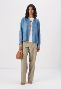 Light blue denim shirt with two chest pockets, white graphic t-shirt underneath, beige wide-leg trousers, and a small brown handbag.