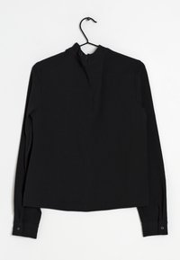 Schwarzes Langarmshirt mit hohem Kragen, ausgestattet mit einem Reißverschluss auf der Rückseite, knöpfbaren Manschetten und einem weichen, strukturierten Stoff. Hängt an einem schwarzen Haken.