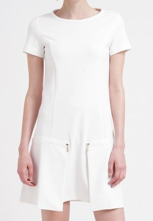 Femme portant une robe blanche à manches courtes avec des poches zippées à l'avant et un col rond sur fond blanc.