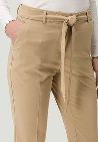 Gros plan sur un pantalon beige avec une ceinture en tissu nouée, poches latérales, porté avec un haut à manches longues blanc, main dans la poche gauche.