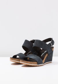 Sandales compensées en suède gris foncé avec des brides croisées, un léger talon en liège et une semelle en caoutchouc. Détail de boucle ajustable au niveau de la cheville.