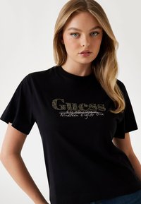 T-shirt nera a maniche corte con logo "Guess" glitterato e testo "Established Nineteen Eighty One". Tessuto di cotone morbido, vestibilità comoda.