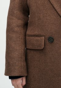 Cappotto in lana a spina di pesce marrone con colletto a risvolto, chiusura a un bottone e tasca laterale con bottone nero.