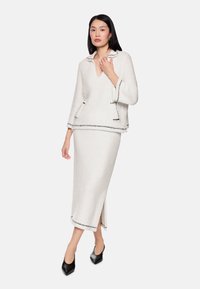 Weißes zweiteiliges Outfit aus strukturiertem Stoff mit einem V-Ausschnitt an der Oberteil und langem Rock, akzentuiert durch schwarze Verzierungen. Das Ensemble umfasst lange Ärmel und einen Seitenschlitz im Rock.