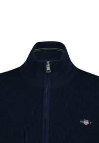 Pull zippé bleu marine côtelé avec col haut et fermeture éclair argentée. Présente un petit logo sur la poitrine, fabriqué en tissu tricoté doux.