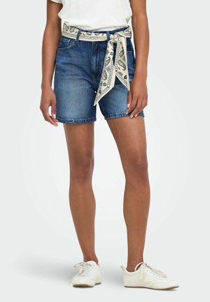 Femme portant un short en denim bleu avec une ceinture foulard blanche à motifs, des baskets blanches et une chemise blanche rentrée.