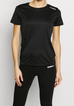 Femme portant un t-shirt de sport noir uni à manches courtes et des leggings noirs avec de petits logos blancs sur l'épaule et la hanche.