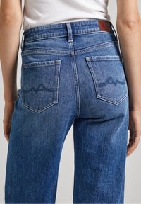 Korkeavyötäröiset siniset farkut, joissa on kaksi takataskua, joissa on hillitty aaltoileva brodeerauskuvio. Kankaan pinta on teksturoidun näköinen ja klassinen malli.