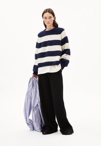 Une femme se tient debout portant un pull rayé bleu marine et blanc, un pantalon large noir, tenant une chemise à rayures bleu et blanc contre un fond blanc uni.
