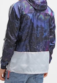 Osoba v pestrobarevném kabátu s galaktickým vzorem a bílou spodní částí, zkombinovaná se světlemodrými džínami.