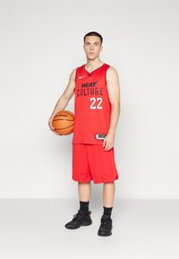 Camiseta de baloncesto roja con "HEAT CULTURE" y el número "22", combinada con pantalones cortos a juego. El jugador sostiene un balón de baloncesto y lleva zapatillas negras.